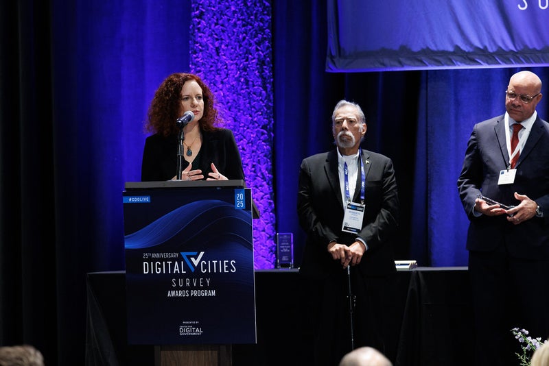 A woman stands at a podium with two men behind her. 